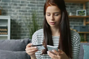 Młoda kobieta siedzi na kanapie, trzyma kartę płatniczą i korzysta ze smartfona.