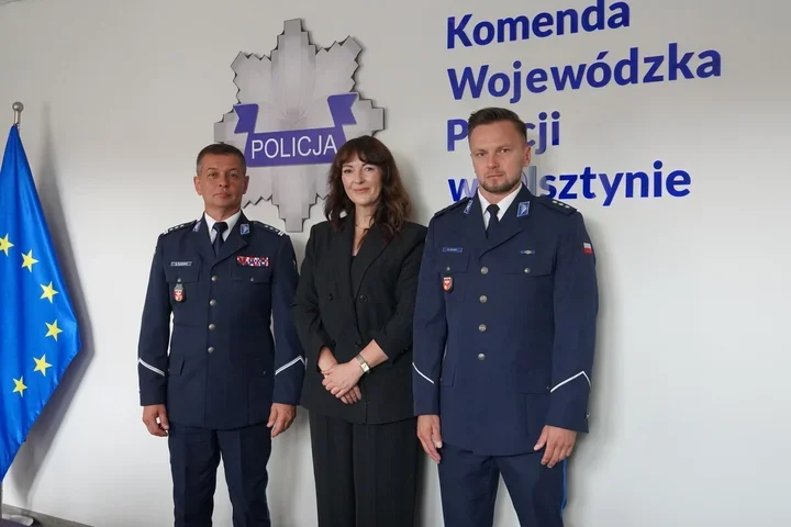 Trzy osoby, w tym dwaj policjanci w mundurach i kobieta w czarnym garniturze, pozują do zdjęcia przed ścianą z logo Policji i napisem Komenda Wojewódzka Policji