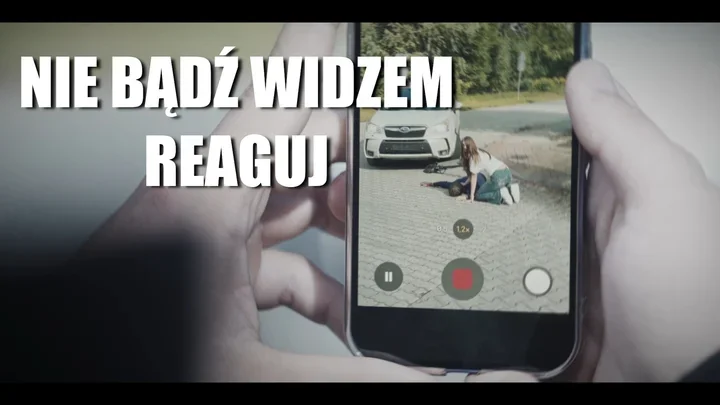 Ręce trzymają smartfon nagrywający wideo, na którym kobieta klęczy przy leżącej osobie na brukowanej nawierzchni przed samochodem; na ekranie hasło "NIE BĄDŹ WI