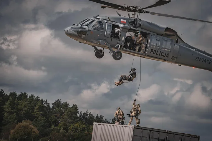 Policyjni antyterroryści ćwiczą zejście na linie z helikoptera Policji na kontener, a dwóch funkcjonariuszy stoi na jego dachu.