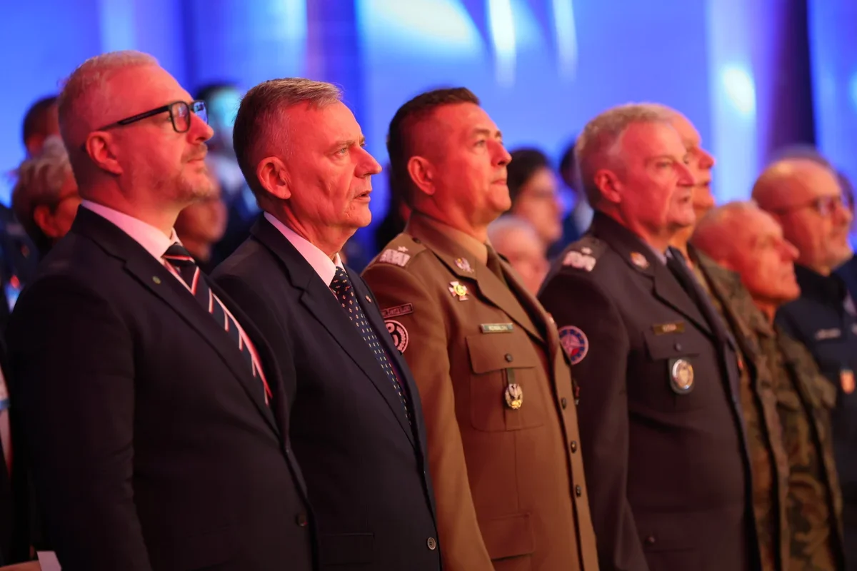 Mężczyźni w garniturach i mundurach wojskowych stoją w rzędzie podczas uroczystości w pomieszczeniu o niebieskim oświetleniu.
