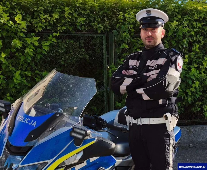 Policjant w mundurze drogówki stoi z założonymi rękami przy policyjnym motocyklu na tle zielonego żywopłotu.