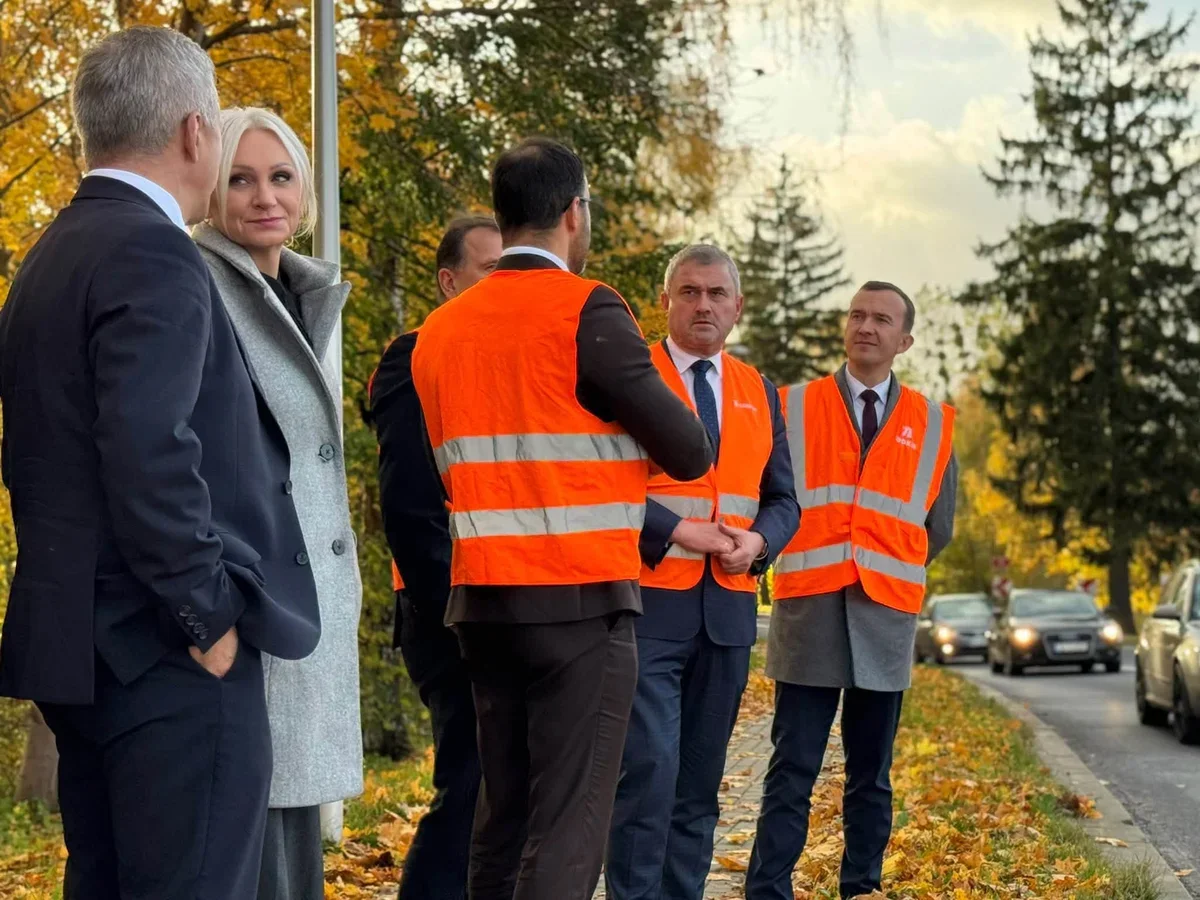 Grupa osób, w tym mężczyźni w pomarańczowych kamizelkach odblaskowych i garniturach oraz kobieta w szarym płaszczu, stoi przy drodze w jesiennym otoczeniu i roz
