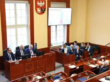 Posiedzenie w sali obrad z udziałem osób w garniturach przy drewnianych biurkach, na ścianie widoczny herb i ekran z prezentacją.