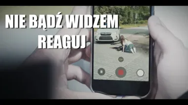 Ręce trzymają smartfon nagrywający wideo, na którym kobieta klęczy przy leżącej osobie na brukowanej nawierzchni przed samochodem; na ekranie hasło "NIE BĄDŹ WI