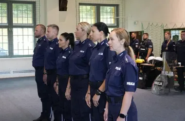 Funkcjonariusze i funkcjonariuszki polskiej policji w mundurach stoją w szeregu w hali sportowej, w tle widoczny sprzęt ochronny i inni policjanci.
