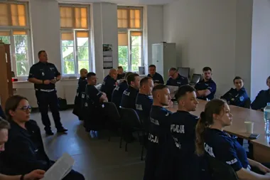 Policjanci w mundurach siedzą przy stołach w sali szkoleniowej, jeden z funkcjonariuszy stoi i przemawia do zebranych.