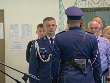 Funkcjonariusz policji w galowym mundurze stoi naprzeciwko innego policjanta w czapce, w tle grupa osób i tablice informacyjne na ścianie.