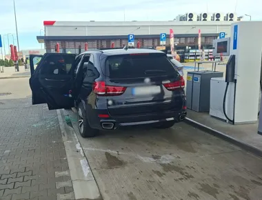 Czarny SUV BMW stoi przy stacji AdBlue na parkingu przed sklepem, z otwartymi drzwiami i rozbitym szkłem na ziemi.