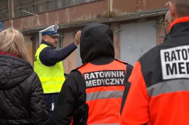 Policjant w kamizelce odblaskowej gestykuluje w kierunku ratowników medycznych stojących przed zabytkowym budynkiem.