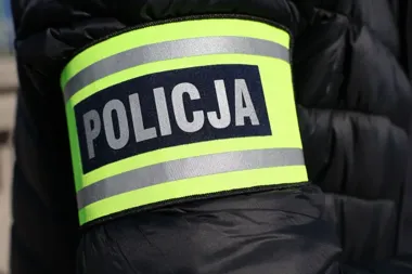 Zbliżenie na żółtą opaskę z napisem POLICJA na ramieniu osoby w czarnej kurtce.