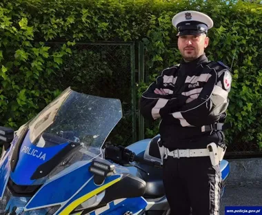 Policjant w mundurze drogówki stoi z założonymi rękami przy policyjnym motocyklu na tle zielonego żywopłotu.