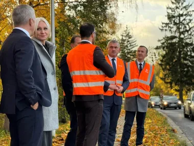 Grupa osób, w tym mężczyźni w pomarańczowych kamizelkach odblaskowych i garniturach oraz kobieta w szarym płaszczu, stoi przy drodze w jesiennym otoczeniu i roz