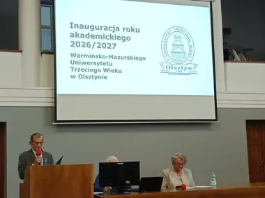 Inauguracja roku akademickiego 2026/2027 Warmińsko-Mazurskiego Uniwersytetu Trzeciego Wieku w Olsztynie w auli, mówca przy pulpicie, dwie osoby przy stoliku.