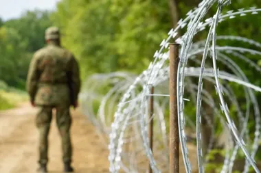 Żołnierz w mundurze kamuflażowym patroluje drogę gruntową wzdłuż ogrodzenia z drutu kolczastego.