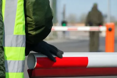 Funkcjonariusz w kamizelce odblaskowej opiera dłoń na zamkniętej szlabanie drogowej na przejściu granicznym.