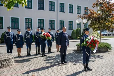 Uroczystość składania wieńców z udziałem funkcjonariuszy służb mundurowych i mężczyzny w garniturze przed budynkiem z zielonymi oknami.