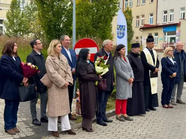 Grupa osób, w tym duchowni i uczestnicy z bukietami kwiatów, stoi w uroczystym szeregu na brukowanej nawierzchni przed budynkiem miejskim.