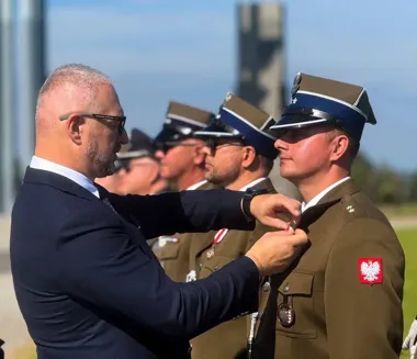 Mężczyzna w garniturze przypina odznaczenie mundurowemu w polskim mundurze wojskowym podczas uroczystości na wolnym powietrzu.
