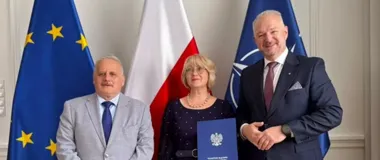 Trzy osoby pozują do zdjęcia przed flagami Unii Europejskiej, Polski i NATO; kobieta w środku trzyma niebieską teczkę z godłem Polski.