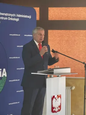 Mężczyzna w garniturze przemawia z mównicy z godłem Polski podczas oficjalnej uroczystości w sali konferencyjnej.