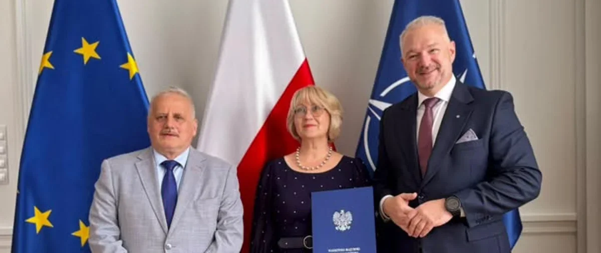 Trzy osoby pozują do zdjęcia przed flagami Unii Europejskiej, Polski i NATO; kobieta w środku trzyma niebieską teczkę z godłem Polski.