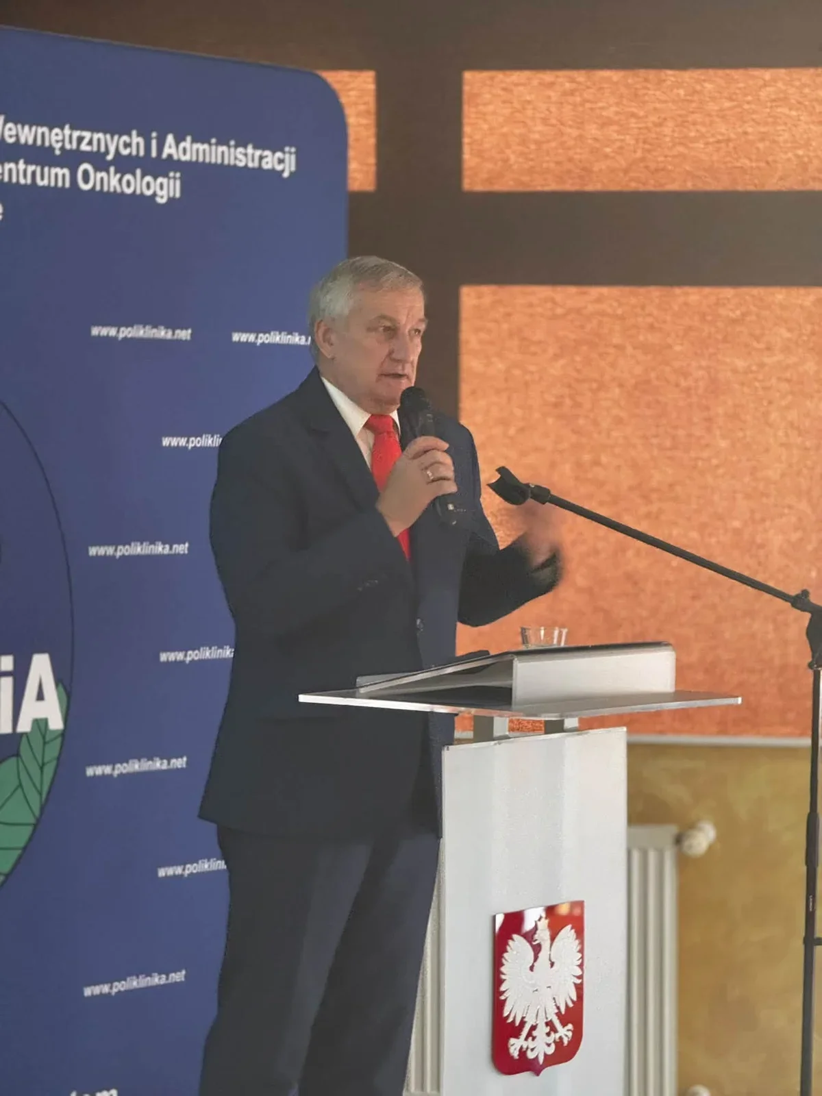 Mężczyzna w garniturze przemawia z mównicy z godłem Polski podczas oficjalnej uroczystości w sali konferencyjnej.